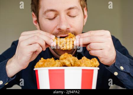 Ragazzo affamato eccitato che mangia le ali di pollo fritte. L'uomo caucasico che gode di cibo spazzatura. Foto Stock