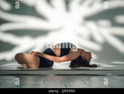 Una giovane donna cacucasiana, che esercita lo yoga. Girato in studio con patterin astratto di luce dietro sullo sfondo. Foto Stock