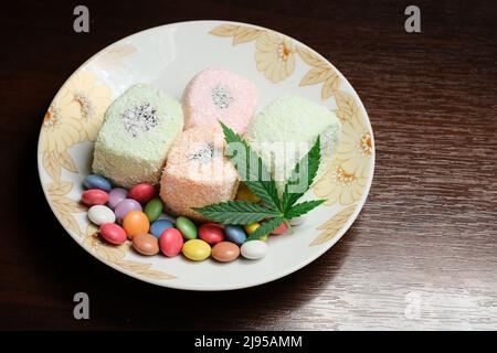 Dessert di cannabis. Delizia turca con fiocchi di cocco e confetti di caramelle colorate, vista dall'alto. Foglie di marijuana verde su fondo piatto bianco. Layout Foto Stock