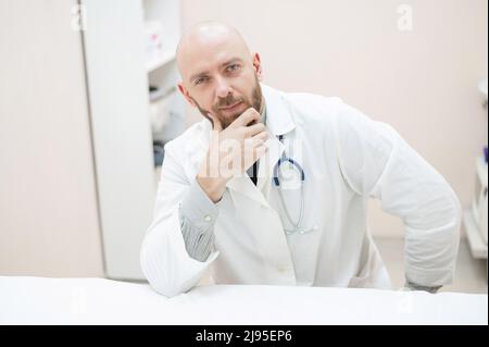 Un medico di sesso maschile si siede ad una scrivania in un ufficio medico. Un calvo con una barba lavora come terapista in una clinica. Cardiologo amichevole con uno stetoscopio Foto Stock