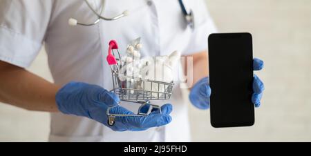 Un medico senza volto in un cappotto medico con uno stetoscopio contiene un mini carrello per lo shopping pieno di diversi medicinali e uno smartphone con uno spazio vuoto Foto Stock