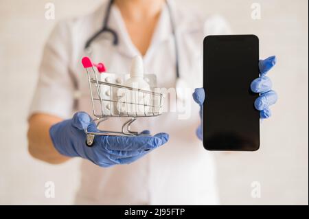 Un medico senza volto in un cappotto medico con uno stetoscopio contiene un mini carrello per lo shopping pieno di diversi medicinali e uno smartphone con uno spazio vuoto Foto Stock