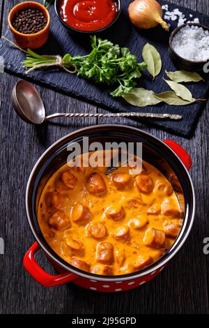 nakkikastike, salsicce finlandesi, si bagna in salsa di pomodoro cremosa in pentola rossa su tavola di legno scuro con ingredienti, orientamento verticale, primo piano Foto Stock