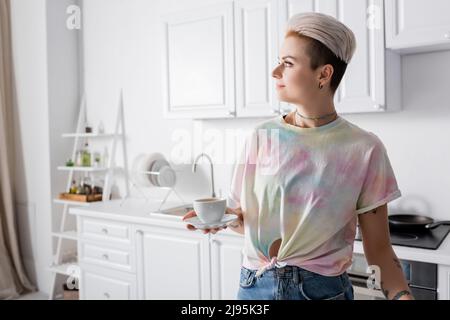 donna con hairstyle trendy che tiene la tazza di caffè e guardando via in cucina Foto Stock