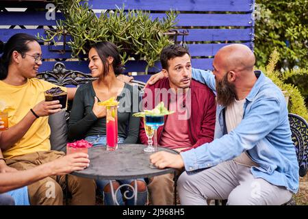 Il gruppo di quattro persone si diverte e ride insieme sulla terrazza del bar, bevendo colorati cocktail di frutta e godendo l'amicizia Foto Stock