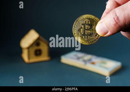 Moneta di Bitcoin in mano sullo sfondo di una casa di legno giocattolo e una pila di cento dollari bollette, fuoco morbido Foto Stock