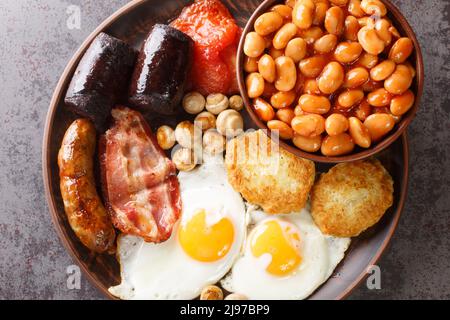 Colazione all'inglese completa con uova fritte, salsicce, pancetta, budino nero, fagioli, pomodoro primo piano nel piatto sul tavolo. Vista orizzontale dall'alto fr Foto Stock