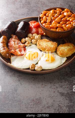 Colazione all'inglese completa con pancetta, salsiccia, uova fritte, fagioli al forno, crocchette di patate e funghi nel piatto sul tavolo. Verticale Foto Stock