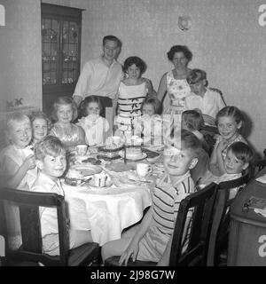1961, storico, compleanno, sorrisi amabili da un gruppo di bambini seduti intorno ad un tavolo godendo di un festeggiamento celebrativo di tè di compleanno, con mamma e papà in piedi vicino per una foto di gruppo, Inghilterra, Regno Unito. Foto Stock