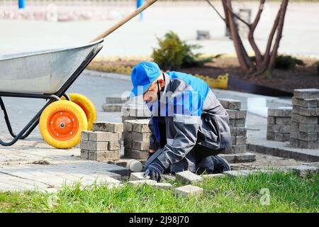 Il muratore in abiti da lavoro si siede sul marciapiede e stende le lastre di pavimentazione. Vista dell'uomo in attività all'aria aperta. Costruttore professionista fa organizzazione del territorio giorno d'estate. Scena reale. Foto Stock