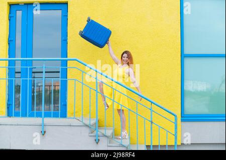 Donna felice con capelli rossi in un abito giallo va al piano superiore tenendo una valigia blu. Foto Stock