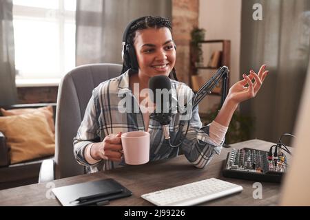 Ritratto medio di giovane donna allegra vestito casual seduta alla scrivania tenendo una tazza di caffè fare contenuto per il video blog Foto Stock