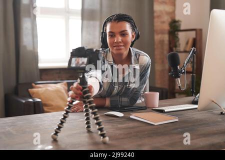 Medio ritratto di bella giovane blogger femmina indossando cuffie seduta alla scrivania in camera loft fissando fotocamera su treppiede Foto Stock