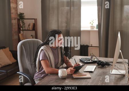 Scatto orizzontale con vista laterale di una giovane donna elegante con trecce afrocapelli vestito casual seduto alla scrivania in camera mansarda utilizzando il pad grafico per cr Foto Stock