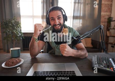 Scatto orizzontale medio di vivace bearded giovane influencer mediorientale che indossa abiti casual seduti alla scrivania da solo godendo qualcosa Foto Stock