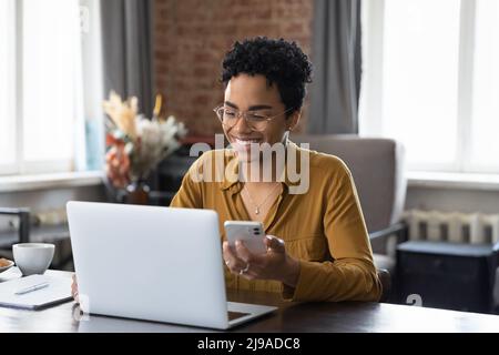 La donna africana si siede alla scrivania tiene il cellulare fissando il laptop Foto Stock