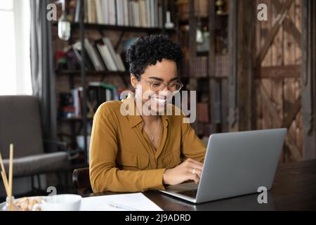 Una donna africana sorridente si siede alla scrivania e lavora su un computer portatile Foto Stock
