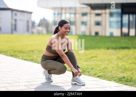 Allegra giovane donna afroamericana che lega i lacci sulle sue sneakers prima dell'addestramento, jogging al parco cittadino, spazio vuoto Foto Stock