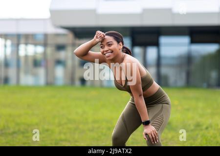 Felice stanca giovane donna nera strofinando sudore dalla fronte dopo il jogging, dopo aver fatto una pausa durante la corsa di allenamento al parco Foto Stock