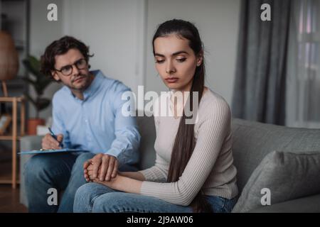 Serio millenario medico europeo in occhiali comforts triste paziente donna seduta sul divano in ufficio Foto Stock