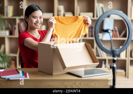 Sorridente signora cinese millennial toglie i vestiti dalla scatola di cartone e guarda la webcam smartphone Foto Stock