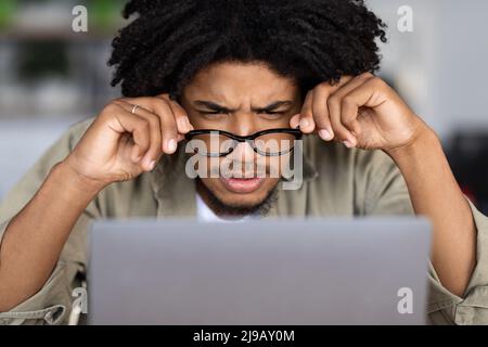 Concentrato curly giovane nero maschio in occhiali guarda lo schermo del laptop al tavolo in caffè interno, primo piano Foto Stock