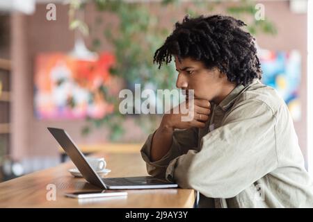 L'uomo nero, giovane e curly, pensivo, guarda lo schermo del computer portatile al tavolo all'interno del bar Foto Stock