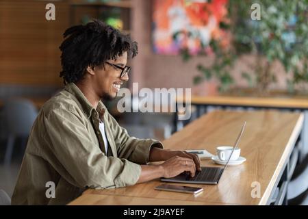 Allegro sorridente ragazzo nero riccio millennial in occhiali digitando sul portatile a tavola in caffè interno, spazio copia Foto Stock