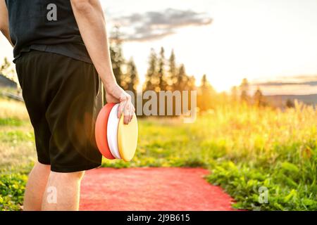 Disco golf in estate al tramonto. Uomo con attrezzatura di frisbee in campo parco. Ragazzo che gioca a discgolf. Giocatore in torneo sportivo all'aperto. Foto Stock