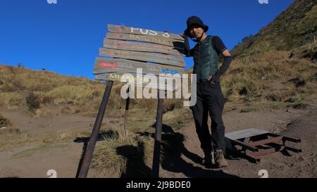 Magelang, Indonesia - 08 06 2019: L'espressione di un uomo in piedi accanto ad un segno mentre si arrampica sul Monte Merbabu su uno sfondo di un chiaro blu sk Foto Stock