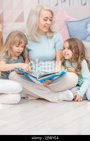 Felice madre bionda che legge un libro alle sue figlie carine che siedono sul pavimento Foto Stock
