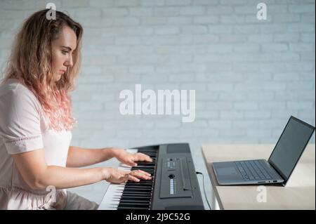 Una donna focalizzata suona il pianoforte elettronico e guarda un corso online sul suo laptop. Musica di apprendimento a distanza messa in quarantena. Rimani a casa. Una lezione in Foto Stock