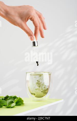 La mano femminile versa la clorofilla liquida in un bicchiere d'acqua con un contagocce. Bicchiere di clorofilla liquida ed erbe fresche sul tavolo verde. Foto Stock