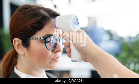 Misurazione della temperatura con termometro elettronico a infrarossi per il personale d'ufficio. Donna infelice con febbre. Un controllo obbligatorio sul lavoro con un termometro Foto Stock