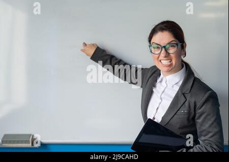 Insegnante arrabbiato in tuta con dito su lavagna bianca in classe. Una professoressa sgraziata tiene un tablet e urla a studenti naughty durante Foto Stock
