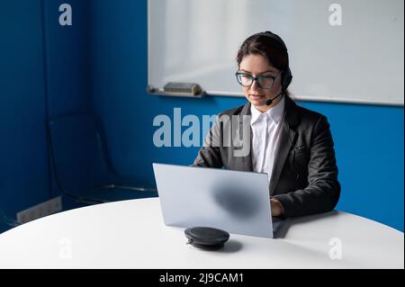 Una donna seria che indossa una cuffia sta conversando online su un laptop in una sala conferenze. Il boss femminile comunica i subordinati tramite videochiamata Foto Stock