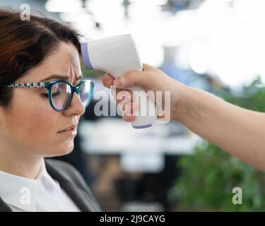 Misurazione della temperatura con termometro elettronico a infrarossi per il personale d'ufficio. Donna malata in un vestito. Il controllo del funzionamento della pistola-termometro è Foto Stock