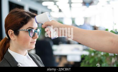 Misurazione della temperatura con termometro elettronico a infrarossi per il personale d'ufficio. Donna malata in un vestito. Il controllo del funzionamento della pistola-termometro è Foto Stock