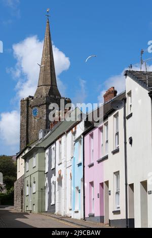 I gabbiani volano dalla torre dell'orologio della chiesa di St Marys e guglia con case color pastello in Cresswell Street alla città portuale di Tenby, Pembrokeshire, Galles Foto Stock