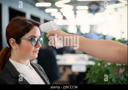 Misurazione della temperatura con termometro elettronico a infrarossi per il personale d'ufficio. Donna malata in un vestito. Il controllo del funzionamento della pistola-termometro è Foto Stock