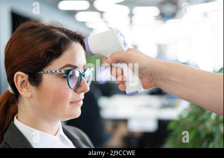 Misurazione della temperatura con termometro elettronico a infrarossi per il personale d'ufficio. Donna malata in un vestito. Il controllo del funzionamento della pistola-termometro è Foto Stock