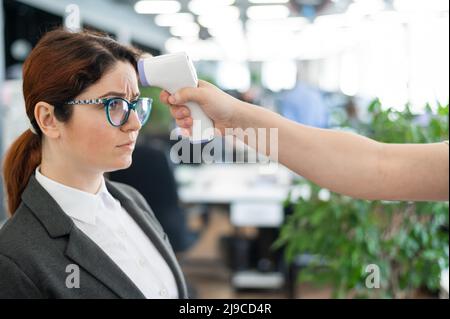 Misurazione della temperatura con termometro elettronico a infrarossi per il personale d'ufficio. Donna infelice con febbre. Un controllo obbligatorio sul lavoro con un termometro Foto Stock