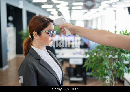 Misurazione della temperatura con termometro elettronico a infrarossi per il personale d'ufficio. Donna malata in un vestito. Il controllo del funzionamento della pistola-termometro è Foto Stock