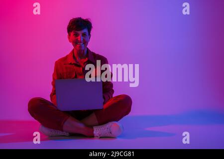 Donna europea matura che lavora con il laptop mentre si siede sul pavimento isolato su sfondo viola Foto Stock