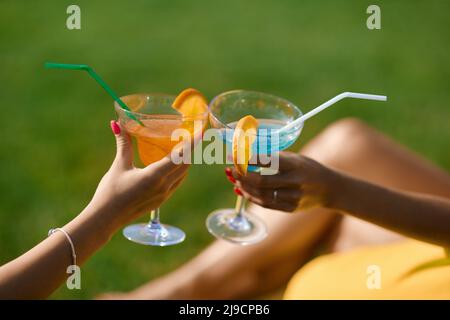 Due mani femminili pulite che si aggraffano con bicchieri di cocktail freschi, con fette di arancia e paillette all'aperto. Vista del raccolto di mani donna curate tostando con bevande, mentre riposano. Concetto di celebrazione. Foto Stock