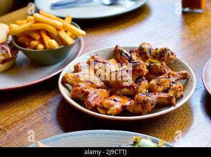 Ali di pollo alla griglia poste sul tavolo da pranzo del ristorante, con patatine fritte (patate fritte) e piatti sfocati sullo sfondo Foto Stock