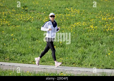 Donna in occhiali da sole e abbigliamento sportivo che corre in un parco con dandelioni fioriti, vista laterale. Solitario runner, allenamento e dimagrimento Foto Stock
