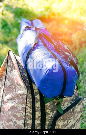 Zaino militare defocus e tenda blu o sacco a pelo. Borsa esercito su sfondo verde erba vicino albero. Zaino militare mimetizzazione. Turismo summ Foto Stock