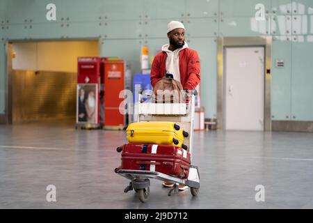 Uomo nero che spinge il carrello dei bagagli a piedi dopo l'arrivo al terminal dell'aeroporto. Vacanza, viaggio. Foto Stock