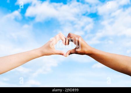 Due mani multietniche si uniscono per formare un cuore di forma con un cielo blu nuvole di sfondo. Concetto di speranza e amore tra razze diverse, pace Foto Stock